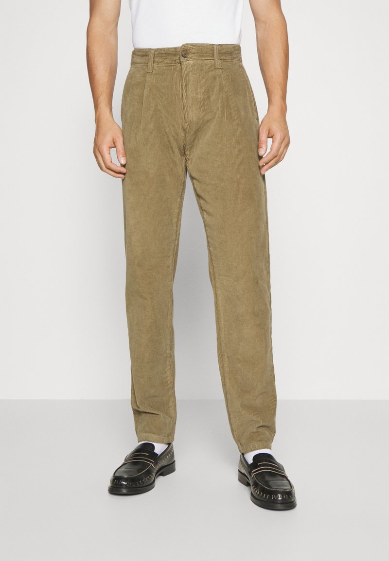 Pantaloni in corduroy verde oliva con design a gamba dritta, dotati di chiusura con bottone, tessuto testurizzato e tasche frontali. Mocassini neri sotto.