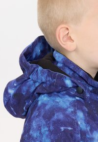 Lilla jakke med galaksemønster, udstyret med hætte og sort kant. Stoffet har en blank overflade med stjerner og nebula-lignende designs.