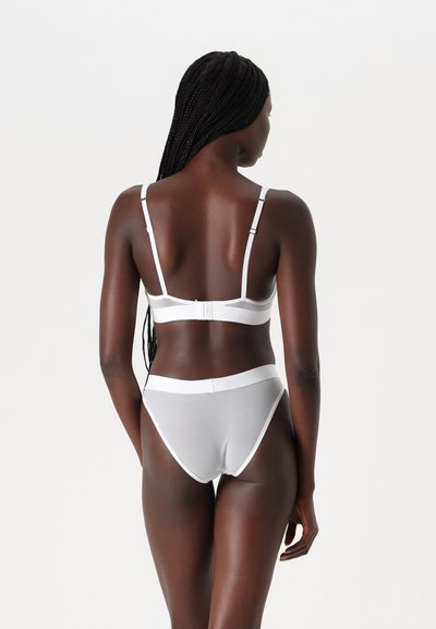 Bela modrčka in visok pas set, izdelan iz mehke tkanine z nastavljivimi naramnicami in elastičnim pasom, ki vsebuje belo obrobo in logotipne detajle.