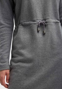Vestido gris de manga larga con cordón en la cintura que presenta terminales metálicos, llevado por una persona con una mano visible a su lado.