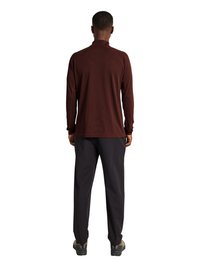 Person stående med ryggen mod kamera iført en langærmet rødbrun top, sorte bukser og vandrestøvler på hvid baggrund.