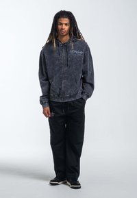 Sudadera con cierre y tie-dye negro, con logo, combinada con pantalones negros de corte holgado. Calzado negro con suelas blancas. Diseño simple y casual.