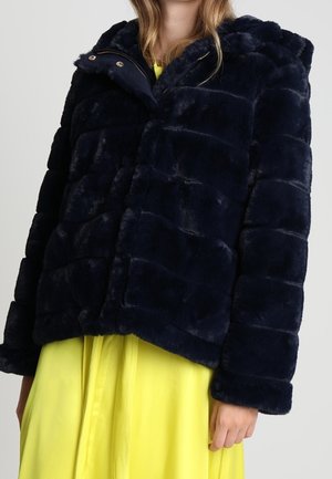 Kobieta ubrana w granatową, sztuczną, puchową kurtkę futrzaną na jasnej żółtej sukience, prezentująca przód i rękawy kurtki.