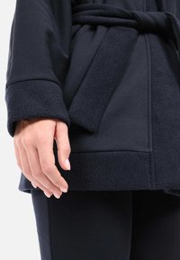 Dunkelblauer Bademantel mit einem weichen Fleecebund und -saum, ausgestattet mit einem passenden Gürtel. Glatte Stoffoberfläche mit einer strukturierten Akzentuierung am unteren Rand.