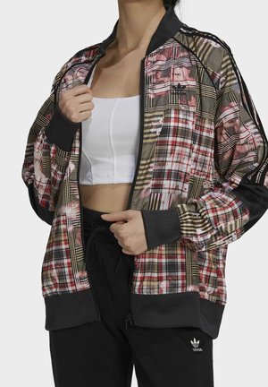 Femme portant une veste zippée à carreaux colorés sur un crop top blanc et un pantalon Adidas noir, ajustant la fermeture éclair de la veste.