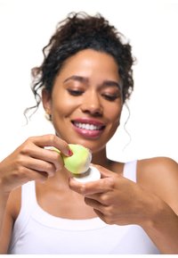 Uśmiechnięta kobieta w białym topie na ramiączkach trzyma zieloną gąbkę do twarzy i białą podkładkę do oczyszczania, przygotowując się do rutyny pielęgnacyjnej.