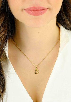 Collier doré avec un pendentif en forme de "d" minuscule, doté d'une chaîne délicate et d'un fermoir simple, porté sur une chemise blanche.