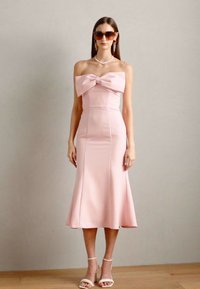 Abito rosa senza spalline con un grande fiocco sul petto, corpetto aderente e orlo svasato. La modella indossa occhiali da sole e tacchi bianchi. Tessuto liscio e opaco.