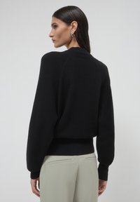 Schwarzer oversized Pullover mit geripptem Saum und Bündchen, rundem Halsausschnitt und weichem Strickstoff. Kombiniert mit hellen Hosen.