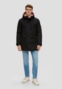 Schwarze Parkajacke mit Kapuze, ausgestattet mit einem grauen und roten Futter, Fronttaschen und einem Reißverschluss. Getragen mit hellblauen Jeans und weißen Sneakers.