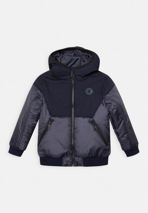 Veste rembourrée navy avec capuche, fermeture éclair, deux poches zippées, poignets côtelés et un logo circulaire sur la poitrine. Matériau lisse et léger.
