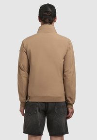 Chaqueta bomber color beige con una textura suave, puños y dobladillo acanalados, y un diseño minimalista. Combinada con pantalones cortos de denim negro, mostrando un ajuste relajado.