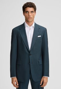 Jonge man in een donkerblauwgroen pak met een wit overhemd en pochet, staand tegen een effen lichtgrijze achtergrond.