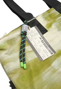 Zielona i kremowa torba materiałowa z teksturowanym wzorem, z czarnym uchwytem oraz kolorowym sznurkiem do ściągania z zielonymi końcówkami i metką podróżną.