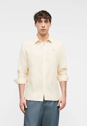 Jonge man die een lichtgele gestreepte lange mouwen shirt draagt met opgerolde mouwen en blauwe broek, staand tegen een effen achtergrond.