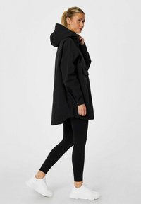 Veste à capuche noire avec une coupe ample, poches latérales et manches à boutons, portée sur des leggings noirs et des baskets blanches.