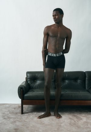 Fiatal férfi fekete Calvin Klein boxeralsóban áll mezítláb a szőnyeges padlón, és egy sötét bőr kanapé mellett pózol, sima fehér fal előtt.