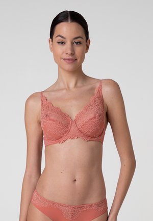 Donna con capelli scuri in reggiseno di pizzo corallo e mutandine abbinate, in piedi davanti a uno sfondo grigio chiaro uniforme.