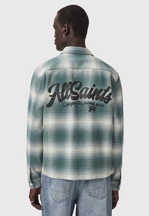 Mand iført grøn og hvid ternet skjorte med teksten "AllSaints Enjoying the journey since 94" på ryggen, kombineret med lyseblå jeans.