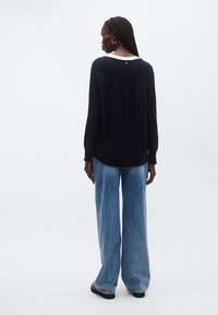 Laivastonsininen neulepaita, jossa on leveä pääntie, pitkät hihat ja pieni yksityiskohta takana. Yhdistetty väljälahkeisiin vaaleansinisiin farkkuihin.