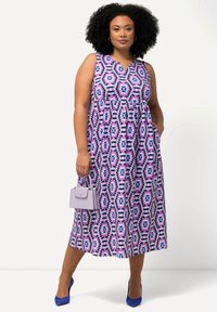 Ärmelloses Midikleid mit Blumenmuster in lila und blauen Tönen, mit V-Ausschnitt, geraffter Taille und Seitentaschen. Begleitet von einer kleinen lila Handtasche.