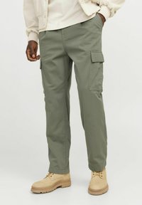 Cargo pants in light olive green fabric, featuring side pockets, straight cut, and worn with tan boots. Smooth texture, casual design.