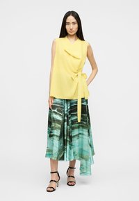 Sieviete valkā bezroku dzeltenu wrap-top ar sānu piesaisti un asimetrisku vidēja garuma svārku ar tirkīzzilu rakstu, kopā ar melniem augstpapēžu sandales.