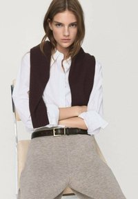 Bruine gebreide trui over een wit overhemd met kraag, grijze broek en een donkere riem; zittende houding tegen een lichte achtergrond.