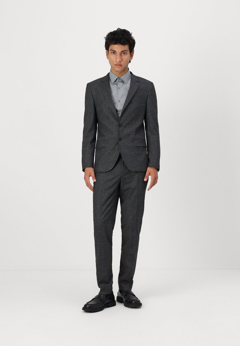 Costume gris charbon avec un motif texturé ; blazer à un bouton, pantalon droit, accompagné d'une chemise habillée gris clair et de chaussures noires.