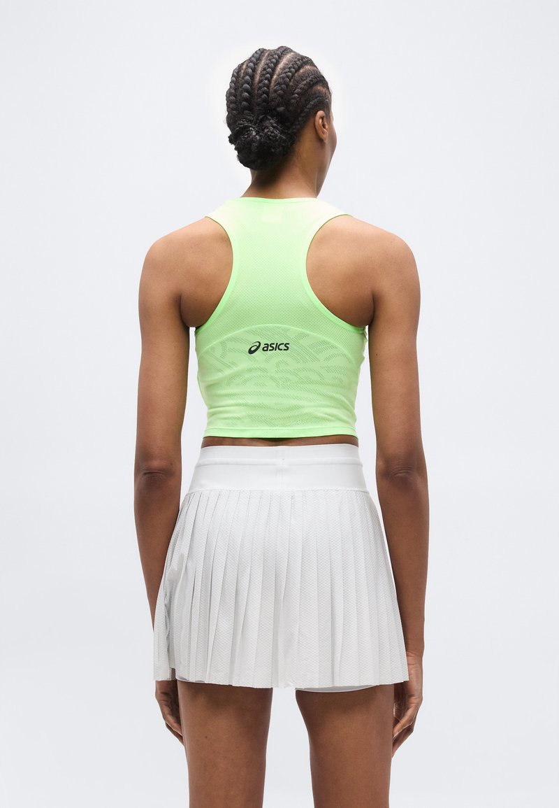 Femme aux cheveux tressés portant un crop top dos nageur vert néon et une jupe plissée blanche, vue de dos sur un fond uni.