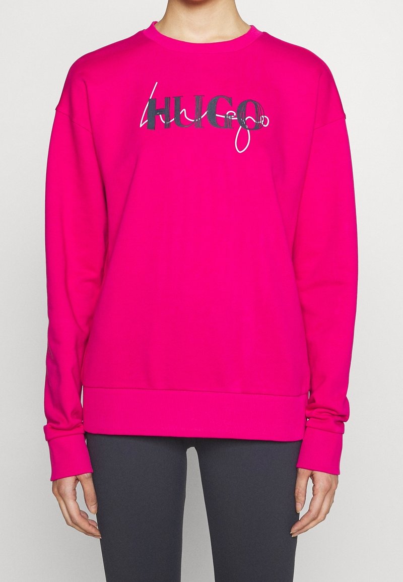 Sweat-shirt à manches longues rose vif avec logo "HUGO" noir et blanc porté par une personne en pantalon noir, debout devant un fond uni clair.