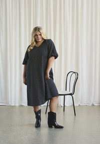Robe noire à pois avec un décolleté en V, des manches amples et un ourlet asymétrique, accompagnée de bottes de cowboy noires sur un fond clair.