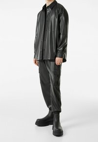 Man med svart konstläderknäppskjorta, svarta cargobyxor med fickor och svarta klumpiga ankelboots, står på vit bakgrund.