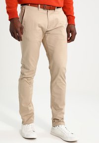 Beige chinos i bomull, med en smal passform och en slät yta, kombinerade med vita sneakers och ett brunt bälte.