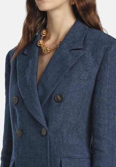 Navy linen double-breasted blazer featuring large brown buttons and notched lapels. Accompanied by a gold textured necklace.
