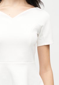 Femme portant une robe blanche à manches courtes avec un col en V et des détails de coutures structurées sur la poitrine.