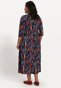 Maxikleid im lebendigen, mehrfarbigen Wellenmuster; lange Ärmel, tailliert, fließender Rock, weicher Stoff. Getragen mit roten Flachschuhen.