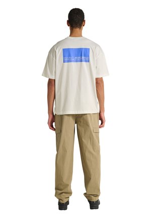 Mężczyzna ubrany w biały, oversize'owy T-shirt z niebieskim prostokątnym nadrukiem i napisem na plecach, beżowe spodnie cargo oraz czarne buty, stoi tyłem.