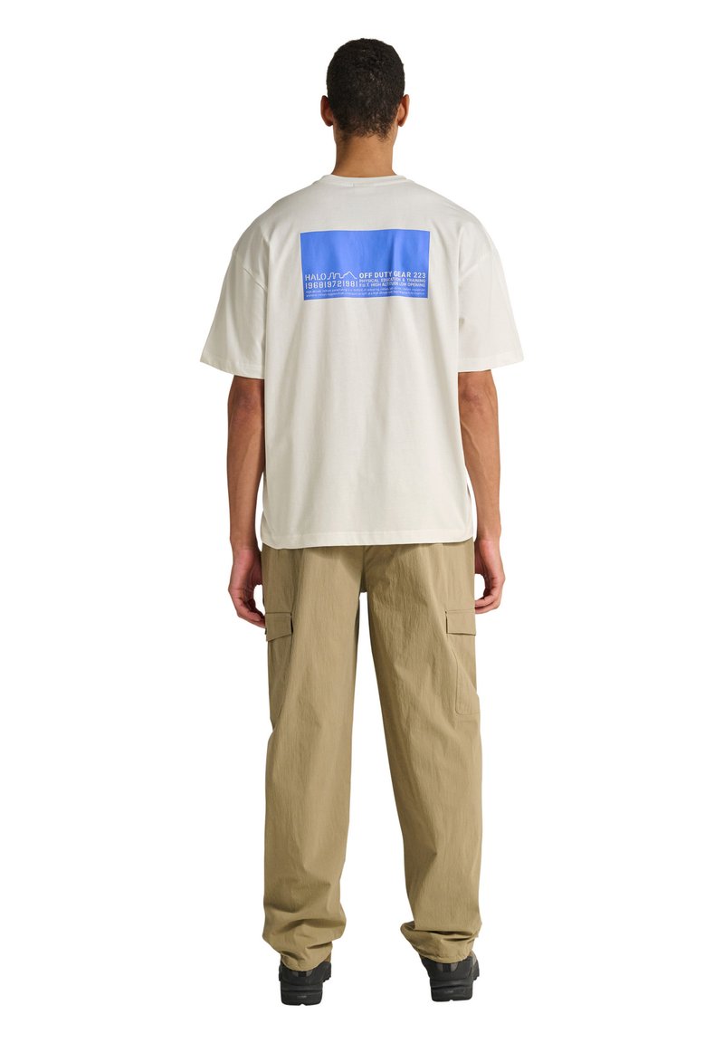 Mann trägt weißes übergroßes T-Shirt mit blauem rechteckigem Grafik- und Textmotiv auf der Rückseite, beige Cargohose und schwarze Schuhe, steht mit dem Rücken zur Kamera.