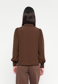 Blusa castanha, texturizada, com colarinho, mangas bufantes e punhos com botões. Usada sobre calças castanhas a condizer, exibida de costas.