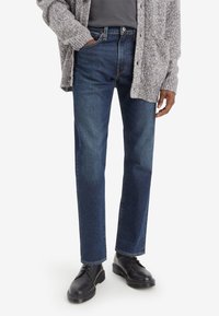 Dark blue denim jeans with a straight fit, featuring belt loops, five-pocket design, and subtle texture details, paired with black shoes.