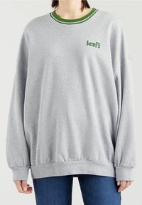 Persona vistiendo un sudadera oversize de color gris claro con ribete verde y el logo de "levis", combinada con jeans azules, frente a un fondo liso.