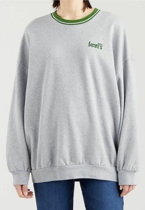 Persona che indossa una felpa grigia chiara oversize con rifiniture verdi e il logo 'levis', abbinata a jeans blu, su uno sfondo semplice.