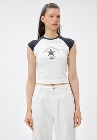 Witte geribbelde crop top met zwarte kapmouwen en bedrukte grafische tekst "Het leven is beter op het strand" met een zeesterontwerp.