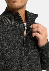 Sort og grå mønstret pullover med høj krave, knaplukning og lynlås. Strikket tekstur med kontrastmaterialer ved kraven.