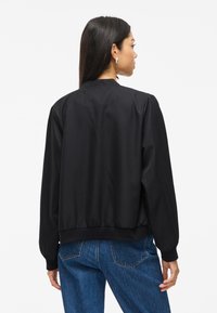 Γυναίκα φορεμένη με μια μαύρη μακριά ζακέτα και μπλε τζιν, στέκεται γυρισμένη προς τα πίσω, δείχνοντας την πίσω όψη της σε άσπρο φόντο.