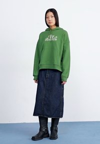 Una persona con lunghi capelli scuri indossa un hoodie verde e una lunga gonna di jeans, abbinata a stivali neri, in piedi di fronte a uno sfondo chiaro.