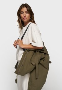 Veste vert olive avec des boutons et des liens, superposée sur une chemise à manches courtes de couleur crème. Possède une sangle noire avec du texte qui traverse l'épaule.