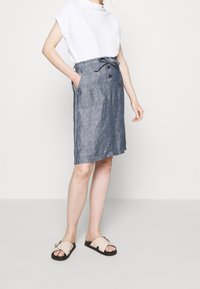 Woman wearing a white sleeveless top, high-waisted blue knee-length skirt with buttons and tie, and beige slide sandals with black soles.