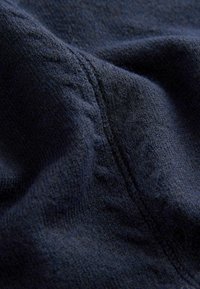 Tmavě modrá látka zblízka, s měkkým, texturovaným povrchem a viditelným švem. Materiál vypadá útulně a pleteně.